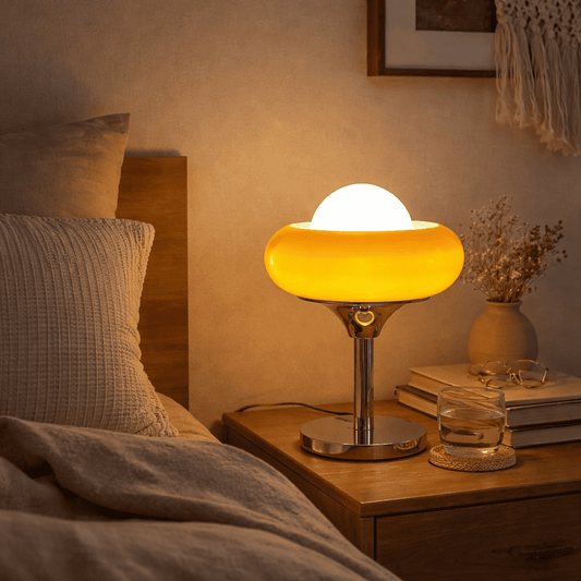 A glowing table lamp with a rounded yellow-orange shade and a white dome top sits on a wooden bedside table