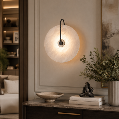 A modern circular wall sconce with a glowing warm white textured disc and a black curved arm