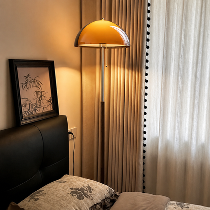 A modern floor lamp with a dome-shaped amber glass shade and a silver metal pole stands beside a bed