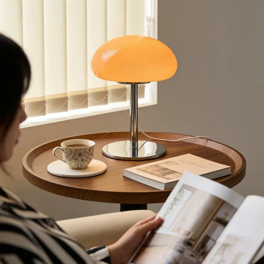 A table lamp with a glowing orange shade and chrome base sits on a round wooden side table