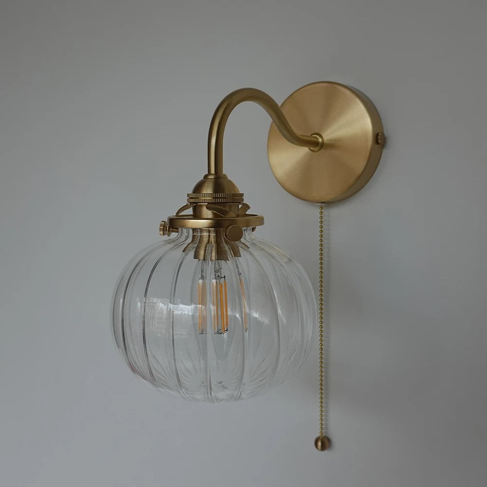 A wall-mounted light fixture with a ribbed clear glass globe shade, an arched brass-toned metal arm