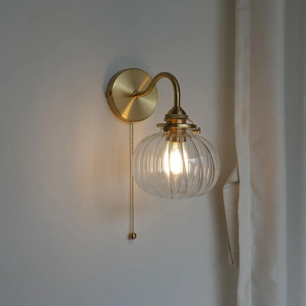 A wall-mounted sconce with a round brass backplate, curved brass arm, and a ribbed clear glass globe shade